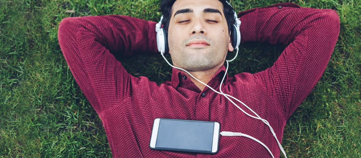 Man lying down in a field while wearing headphones plugged into a smartphone