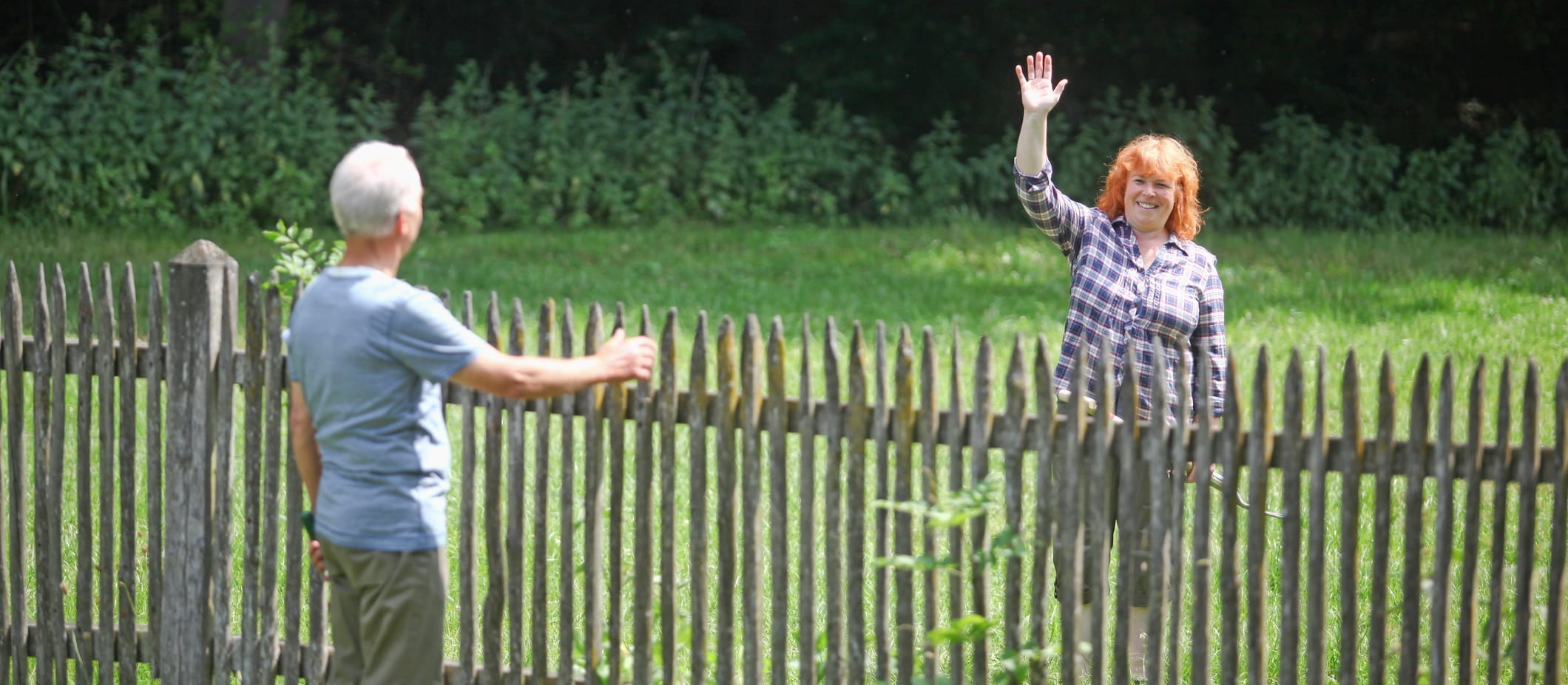 two neighbours waving form across a fence