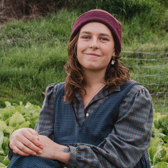 Headshot of Libby who has shoulder length brown hair, a beanie on and a dress over a checkered long sleeve shirt. 