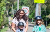 A mother stands next to her two young children, one is holding a bike and the other a helmet. They are all smiling looking at the camera. 