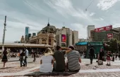 Three young adults sit facing Flinders Street Station. Thier backs are to camera.