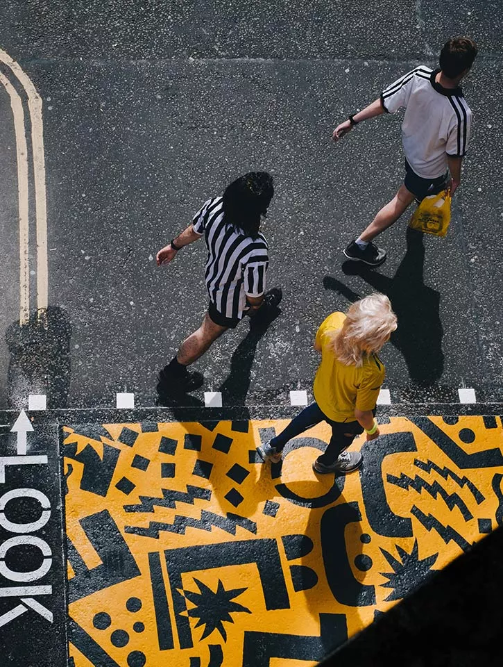 Birds eye view of three people walking together. The ground has been painted with a yellow and black artwork. They people are dressed to match.
