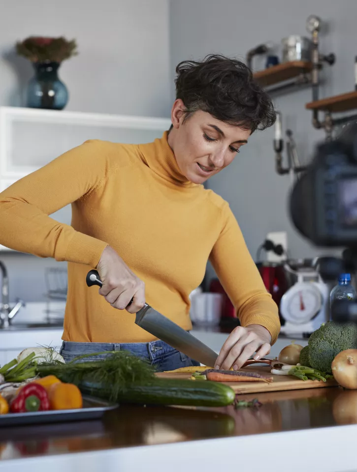 A woman wearing a yellow turtleneck is chopping up vegetables. on the bench next to her is a baking tray of vegetables. 
