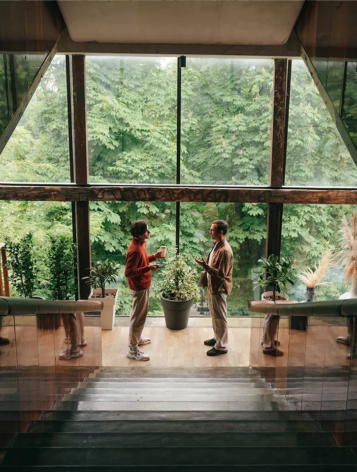 Two men are standing at the bottom of the stairs, they are having a conversation. Behind them is a huge floor to ceiling window looking out on beautiful greenery. 