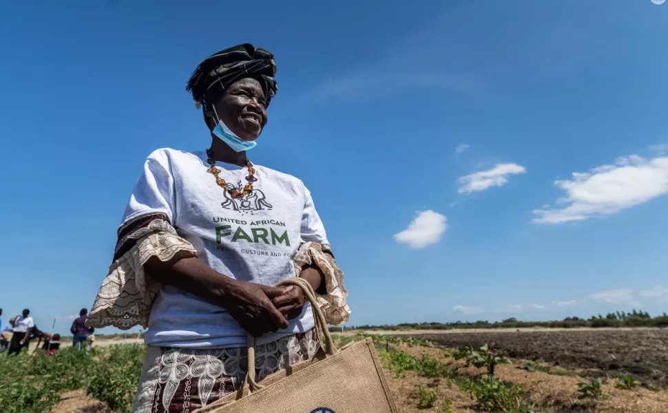 A woman wearing a United African Farm t-shirt and bag is standing in a garden.  The sky is a beautiful blue. 